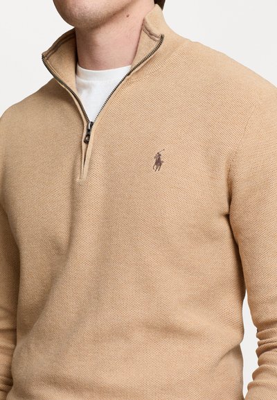 Sweater bege com fecho éclair em malha texturizada, apresentando um pequeno logo no lado esquerdo do peito e uma camada interior branca visível na gola.