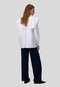 Donna con lunghi capelli rossi, indossa una camicia bianca con un grande colletto ricamato e pantaloni jeans blu scuro a gamba larga, in piedi di lato su uno sfondo grigio.