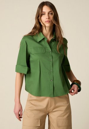 Kvinde iført en grøn skjorte med knapper med opkrøllede ærmer og beige cargobukser, accessoriseret med mørke armbånd på højre håndled.