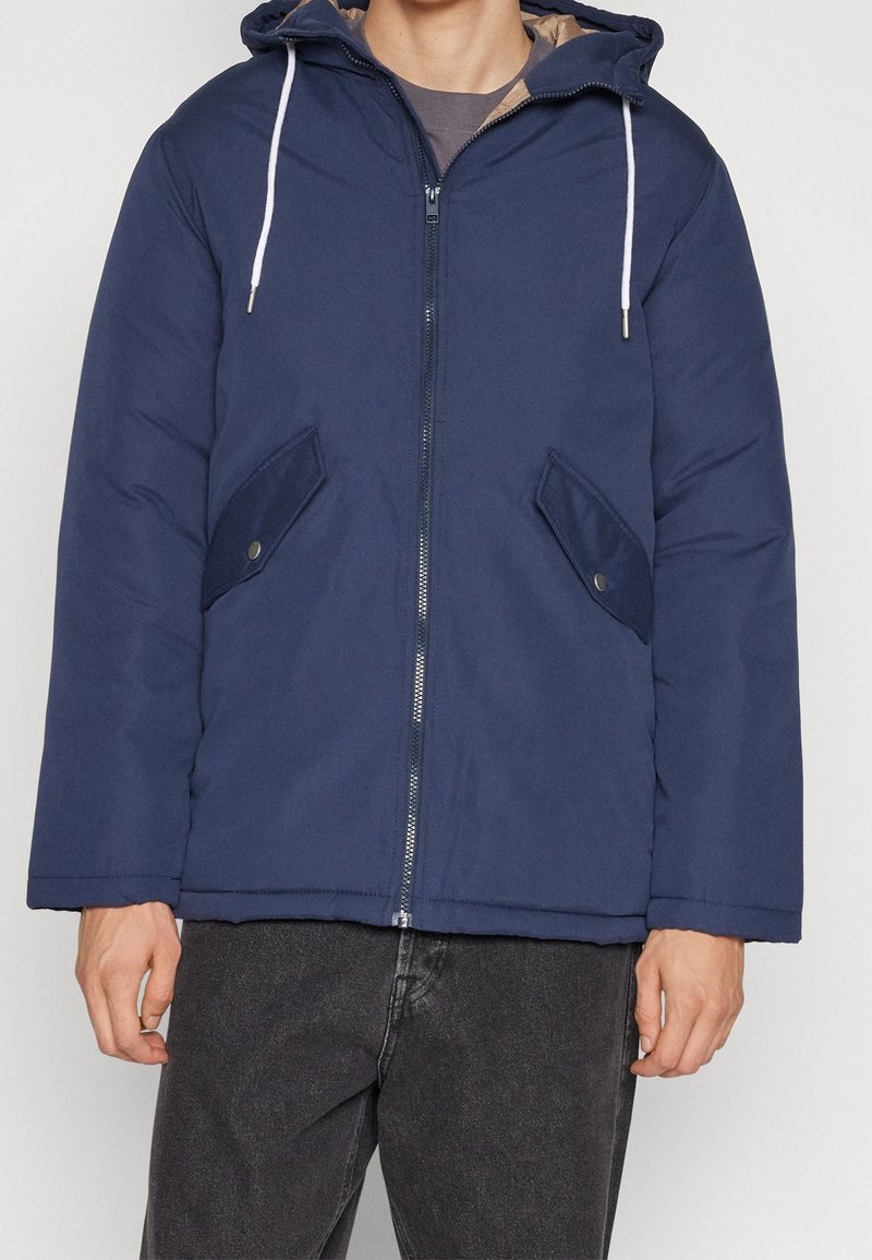 Giubbotto imbottito blu navy con zip frontale, due tasche laterali e cappuccio regolabile con cordino. Presenta un'esterno liscio e un rivestimento morbido.
