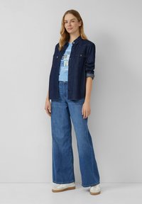 Chemise en denim bleu foncé, style boutonné, avec deux poches poitrine. En dessous, un t-shirt graphique bleu clair. Jean large bleu et baskets blanches.