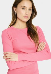 Roze top met lange mouwen en ribbels, met een ronde halslijn, gedragen door een persoon met gekruiste armen. Gelaagd stof, geen zichtbare patronen of logo's.