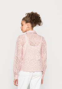 Blusa rosa de eyelet com bainha em ponta, mangas longas e recortes florais, apresentando um colarinho e punhos com botão.