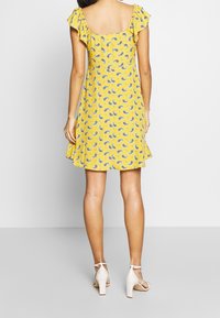 Keltainen mekko paisley-kuvialla, lyhyet röyhelöhihat, neliöselkä ja levenevä helma. Mallilla on päällään valkoiset paksupohjaiset sandaalit.