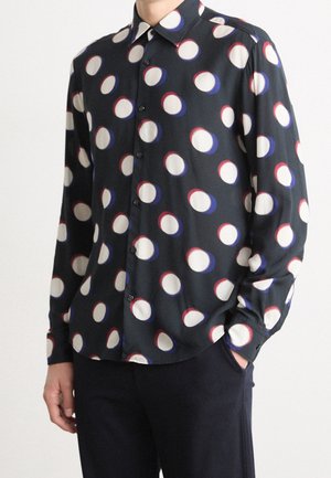 Homme portant une chemise noire à boutons avec de grands cercles blancs bordés de rouge et de bleu, associée à un pantalon noir.