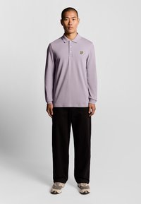 Polo a maniche lunghe di colore viola chiaro con colletto e logo farfalla gialla ricamato, abbinato a pantaloni neri a gamba larga e sneaker beige.