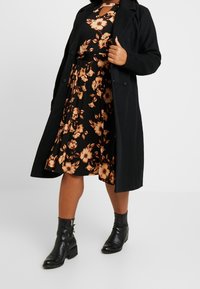 Abrigo negro sobre un vestido floral con flores naranjas. El vestido tiene una cintura ceñida y una falda acampanada. Botines negros con detalle de cremallera.