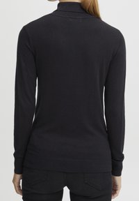 Frau von hinten gesehen, die einen schlichten schwarzen Langarmpullover und schwarze Jeans trägt, mit im Nacken gebundenem Haar zu einem tiefen Pferdeschwanz.