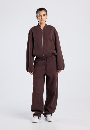 Bruine trui met een ritssluiting aan de voorkant en een hoge kraag, gecombineerd met bruine joggingbroek met een elastische tailleband, gestyled met witte sneakers.