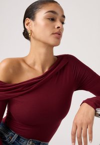 Μπουρντό τοπ με έξω τους ώμους από απαλό ύφασμα με draped λεπτομέρειες. Μακριά μανίκια και εφαρμοστή γραμμή. Το μοντέλο φοράει τζιν με λεπτή γραμμή.