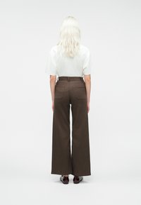 Person med langt blondt hår står med ryggen vendt, iført en hvid kortærmet skjorte, høj talje, vide brune bukser og mørke sko på hvid baggrund.