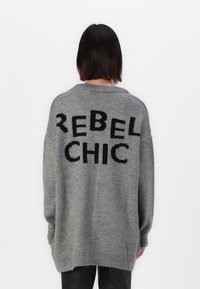 Suéter gris de gran tamaño hecho de material suave, con un gran texto negro "REBEL CHIC" en la parte posterior y puños acanalados.