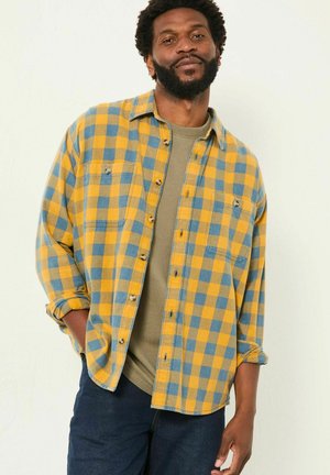Gelb und blau karierte Flanellhemd mit Knopfverschluss und Brusttaschen, über einem olivgrünen Pullover getragen. Dunkle Jeans.