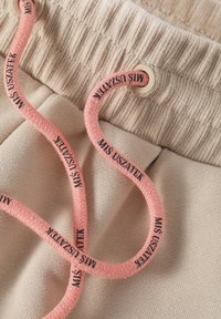 Beiga mjukisbyxor med ribbad midja och en rosa dragsnodd med svart text, lagda på en texturerad yta.