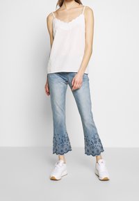 Haut blanc sans manches avec un col festonné, associé à un jean bleu clair avec des motifs floraux brodés au bas et des baskets blanches.