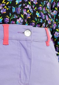 Liliowe spodnie z różowym przeszyciem i guzikiem, o gładkiej fakturze. Czarna kwiecista bluzka z fioletowo-zielonymi akcentami na górze.