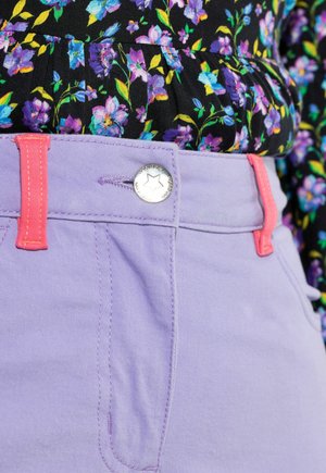 Liliowe spodnie z różowym przeszyciem i guzikiem, o gładkiej fakturze. Czarna kwiecista bluzka z fioletowo-zielonymi akcentami na górze.