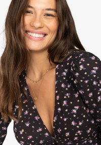 Rochie neagră cu flori, cu mâneci bufante, prezentând flori mici roz și albastre, asortată cu coliere aurii suprapuse și un decolteu adânc.