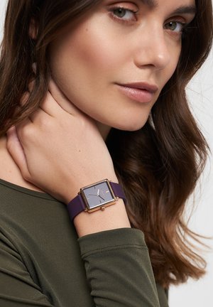 Square watch with a gray face, gold accents, and a purple silicone strap, worn on a wrist with a dark green top.