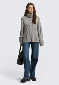 Grijze kabelgebreide turtleneck trui met geribde zoom, gecombineerd met uitlopende blauwe jeans en een zwarte handtas. Chunky zwarte schoenen ronden de outfit af.