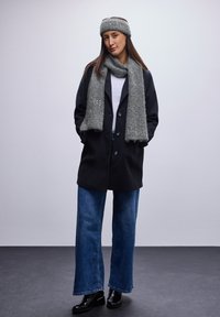 Cappotto nero, sciarpa grigia e fascia per capelli, t-shirt bianca, jeans blu a gamba larga, stivaletti neri alla caviglia con dettagli dorati, texture di tessuto morbido.