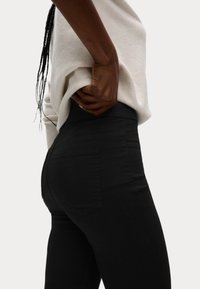 Jeans negros ajustados con cintura alta, que cuentan con bolsillos traseros. Llevados con un suéter color crema, resaltando el diseño estilizado y la textura de la tela.