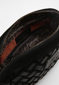 Musta kudottu mokkasaumabag, jossa on vetoketjulla avattava suu, paljastaa sileän sisäosan, joka on vuorattu kuvioidulla kankaalla ja nahkakoristeella.