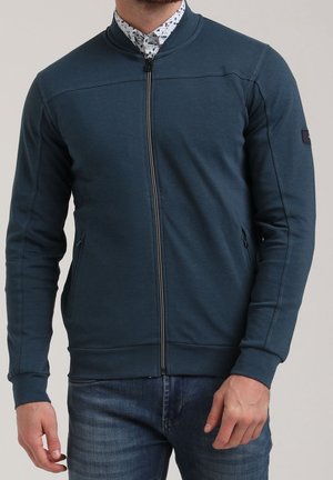 Mężczyzna ubrany w ciemnoturkusową kurtkę na zamek błyskawiczny, pod którą ma wzorzystą białą koszulę, w niebieskich dżinsach, stojący na jednolitym tle.