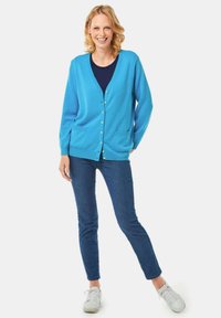 Hellblauer Cardigan mit Knöpfen, zwei vorderen Taschen, über einem dunkelblauen Oberteil getragen, kombiniert mit dunklen Jeans und weißen Sneakers.