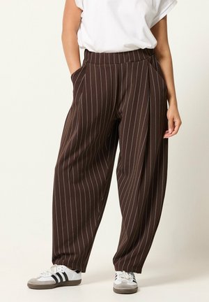 Femme portant un pantalon ample marron à fines rayures avec des poches et des baskets blanches, associé à un t-shirt blanc à manches courtes.