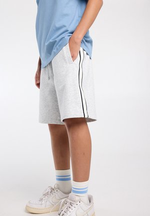 Persoon draagt lichtgrijze korte broek met zwart-witte strepen, blauw T-shirt, witte sneakers en witte sokken met blauwe strepen, staat zijwaarts.
