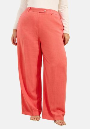 Frau trägt korallfarbene Weitbein-Hose mit Knopf und Gürtelschlaufen, kombiniert mit beigen offenen Sandalen und einem weißen Langarm-Top.