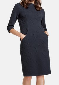 Abito blu navy con maniche a tre quarti, tessuto testurizzato, tasche laterali e una silhouette aderente fino al ginocchio.
