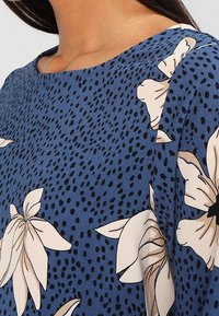 Haut floral bleu présentant un tissu texturé avec un motif de grandes fleurs crème et des points noirs, encolure arrondie et coupe ample.