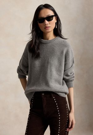 Kvinde med langt mørkt hår iført sorte solbriller, grå strikket sweater og mørke bukser med sølvnitter, der står mod en ensfarvet beige baggrund.
