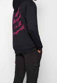 Svart huvtröja med rosa grafisk text på baksidan. Tillverkad av mjukt tyg med ribbade ärmslut och nederkant, i kombination med svarta cargobyxor.