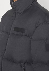 Czarna puchowa kurtka z wysokim kołnierzem, pikowana, z zapięciem na zamek i naszywką z logo. Miękki, ocieplany materiał z akcentem przy zamku.