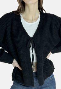 Schwarzer gestrickter Cardigan mit V-Ausschnitt und mittigem Bindebandverschluss. Mit lockeren Ärmeln und einem strukturierten Muster. Über einem weißen Oberteil getragen.