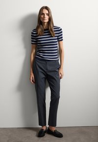 T-shirt a righe navy e bianche con maniche corte abbinato a pantaloni scuri grigio su misura e mocassini neri, posizionato davanti a un muro semplice.