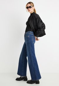 Μαύρο μπουφάν bomber με σούρες στα μανίκια, μαύρο εφαρμοστό τοπ, μπλε τζιν παντελόνι με φαρδιά εφαρμογή και μαύρα μποτάκια μέχρι τον αστράγαλο. Τα γυαλιά ηλίου ολοκληρώνουν την εμφάνιση.