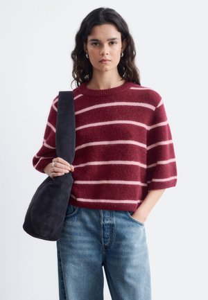 Jonge vrouw draagt een bordeauxrood en roze gestreepte trui en blauwe jeans, houdt een zwarte schoudertas vast met één hand in haar zak.