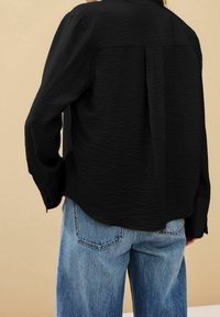 Blusa nera in tessuto testurizzato con una vestibilità rilassata, caratterizzata da una piega sulla schiena, maniche lunghe e abbinata a jeans blu a vita alta.