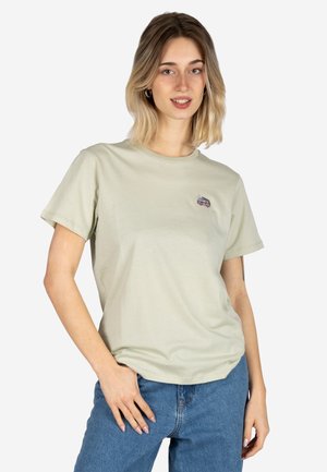 Young woman with shoulder-length blond hair wearing a light beige t-shirt with a small camper van patch and blue jeans.