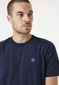 Camiseta de cuello redondo azul marino hecha de una tela texturizada, con un pequeño logo cuadrado en el área del pecho izquierdo. Mangas cortas, corte relajado.