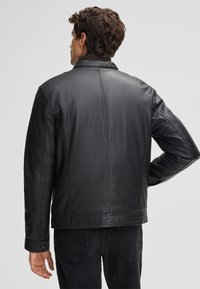 Giacca di pelle nera con una texture liscia, dotata di colletto, design aderente e dettagli di cucitura visibili, indossata sopra un jeans scuro.