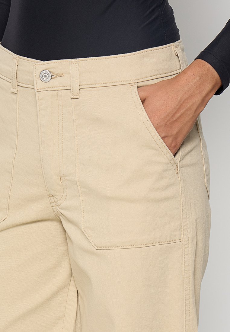 Mano descansando dentro del bolsillo delantero de unos pantalones beige, combinados con una camiseta de manga larga negra, mostrando costuras detalladas y un cierre de botón metálico.