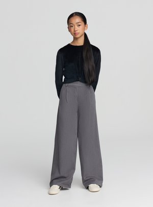 Czarna aksamitna bluzka z długim rękawem i wysokie, szare spodnie o szerokich nogawkach z delikatną fakturą, zestawione z białymi trampkami.