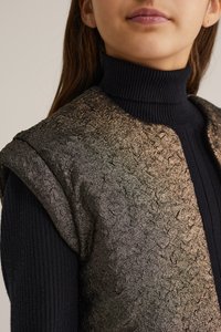 Skinande, texturerad väst med en mörk, iriserande finish, kombinerad med en figurnära svart ribbad polotröja. Närbild av axel- och halsringningsdetaljer.
