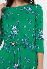 Zelené květované šaty zdobené různými modrými a bílými květinami. Design doplňují tříčtvrteční rukávy a zavázání v pase.