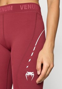 Leggings atléticas cor de castanha com uma cintura larga, apresentando logótipo branco e elementos de design. Tecido suave com brilho subtil e elasticidade.